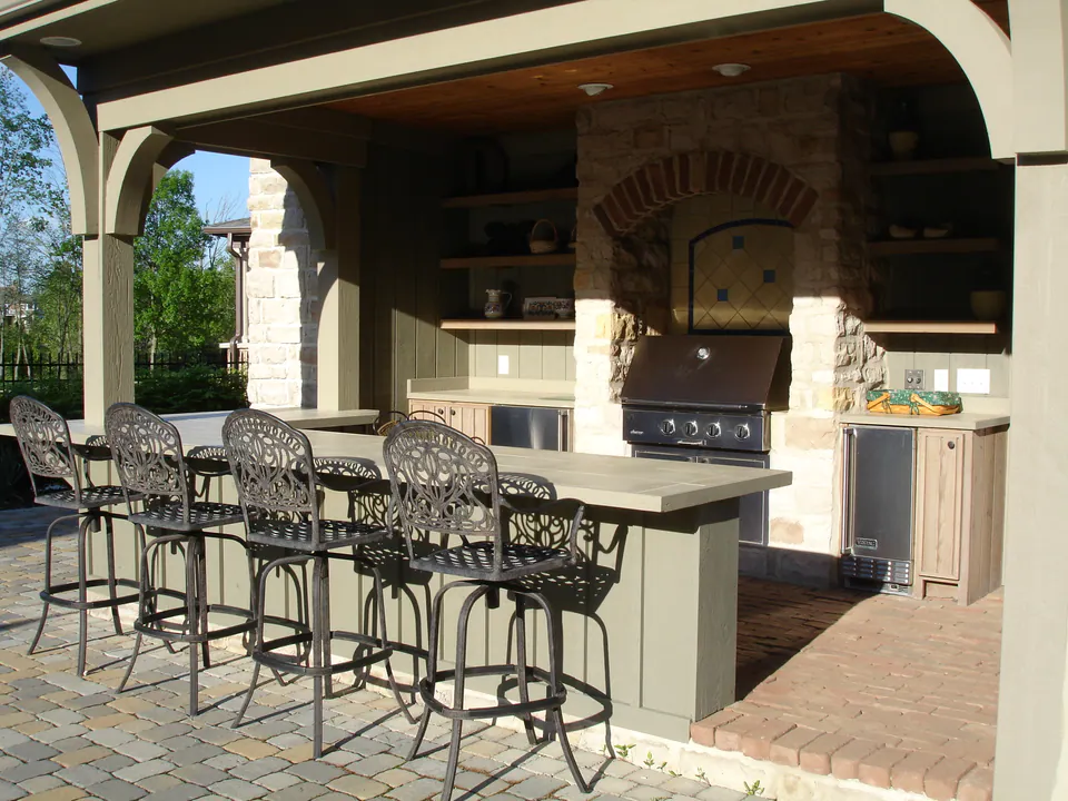 Covered outdoor kitchen with a stone pizza oven, grill, bar counter, and wrought-iron stools under a wood ceiling